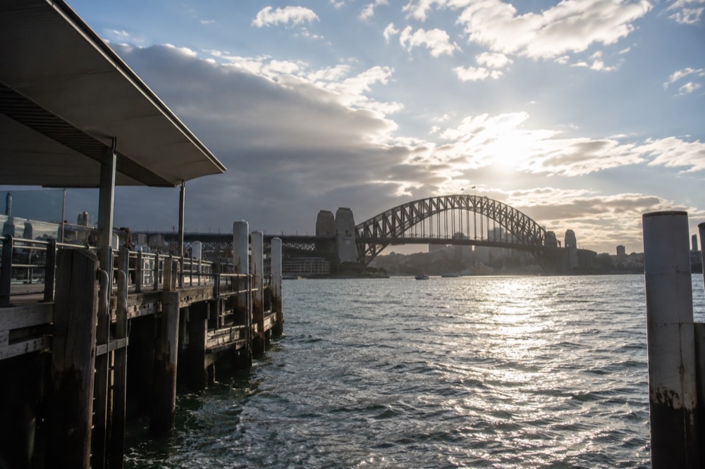The arch of a steel bridge rising above harbour water with the city skyline beyond