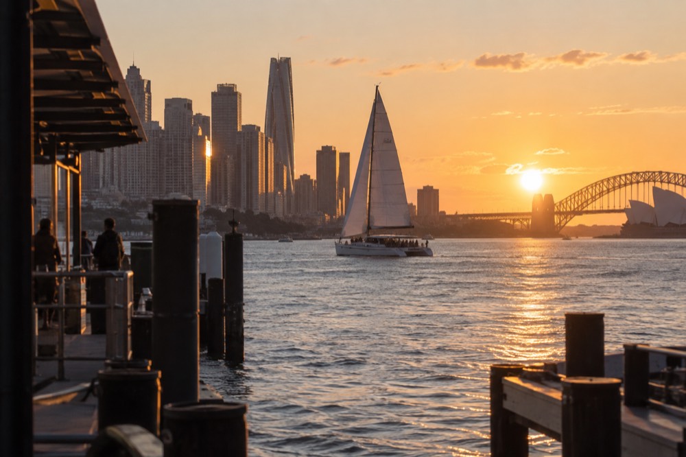 A catamaran under sail at sunset with city towers glowing behind it