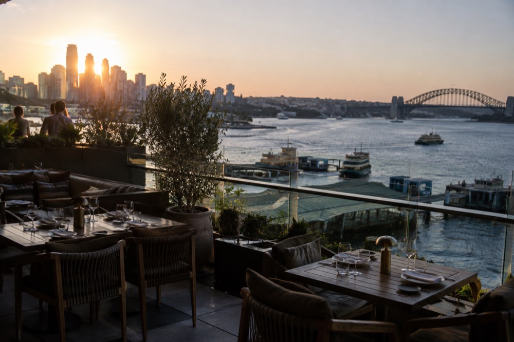 A rooftop bar at sunset with the Sydney skyline and harbour in the background, empty setting tables