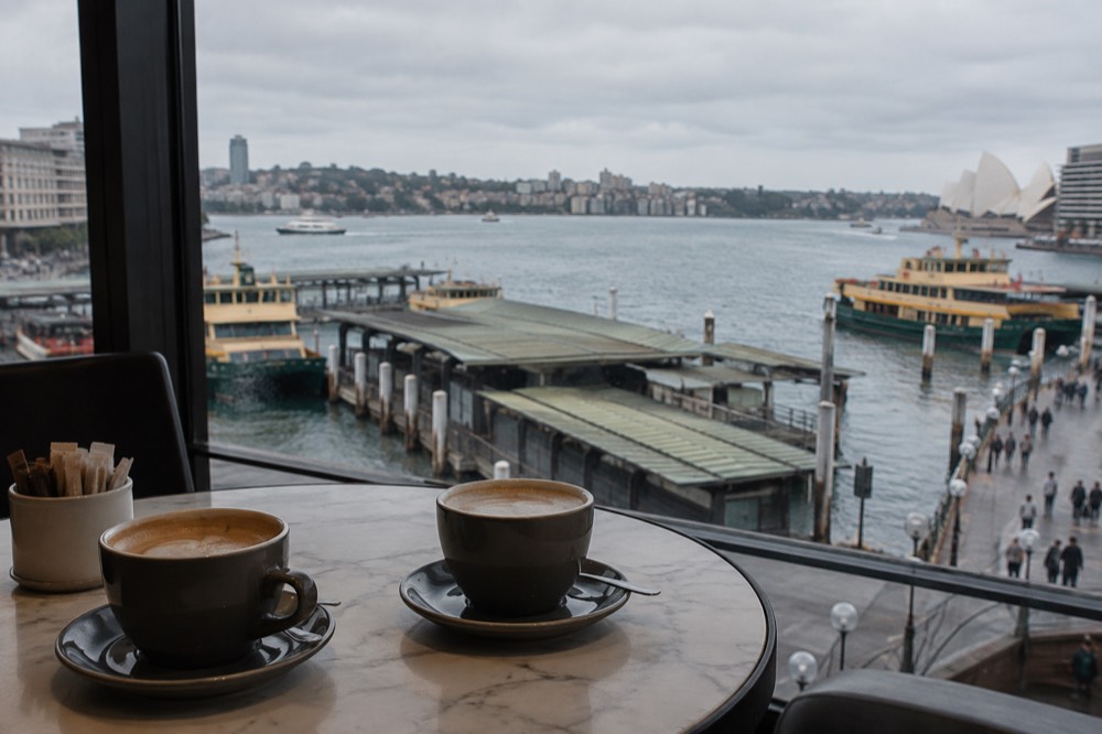 Two coffee cups on a small café table beside a window with harbour water outside
