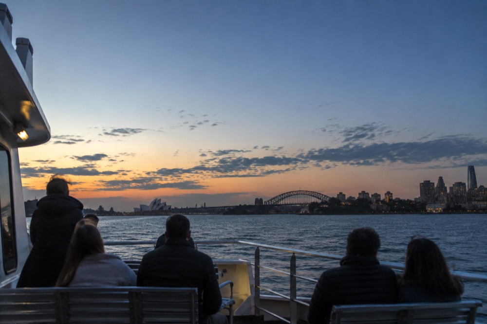 Passengers on a catamaran deck watching the sun set over Sydney Harbour