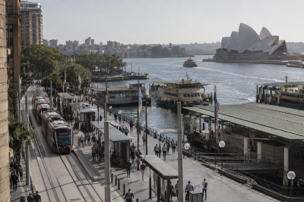 A light rail stop platform in central Sydney with a tram approaching and heritage architecture behind
