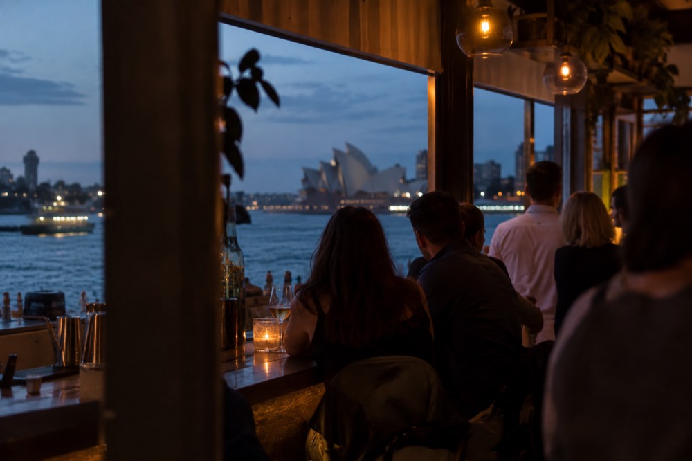 A warmly lit small bar interior with people gathered along a timber counter in the evening