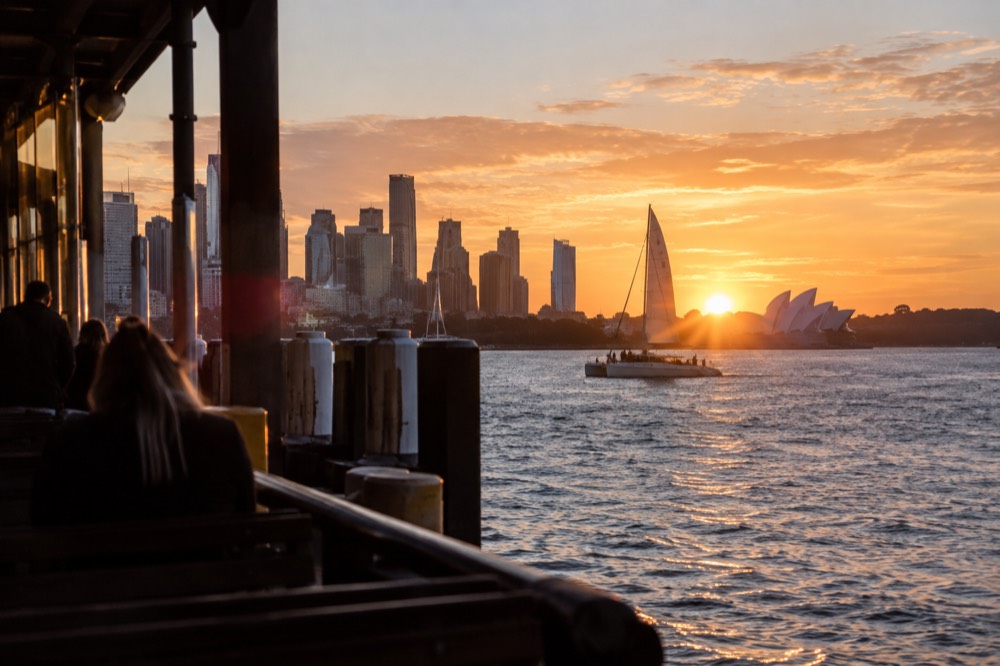 A catamaran under sail on the harbour with the city skyline glowing orange at sunset