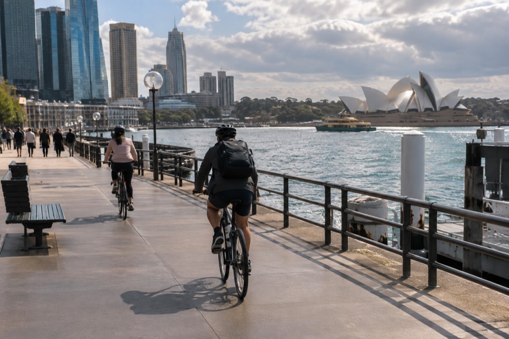 Cyclists on a wide flat path beside harbour water with city skyline