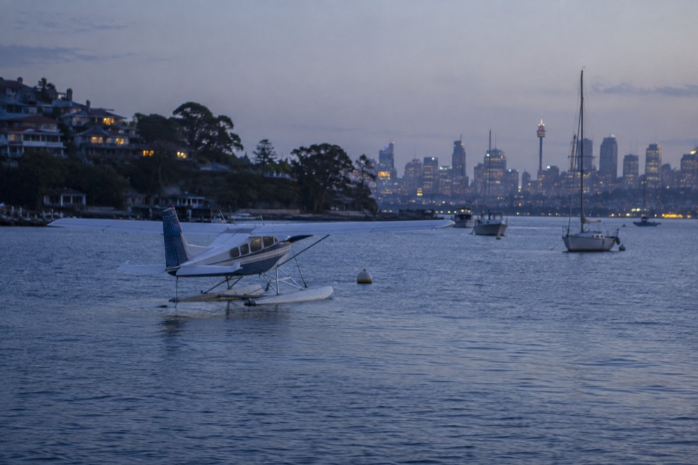 A light seaplane floating on the calm water of Rose Bay with Sydney Harbour in the background