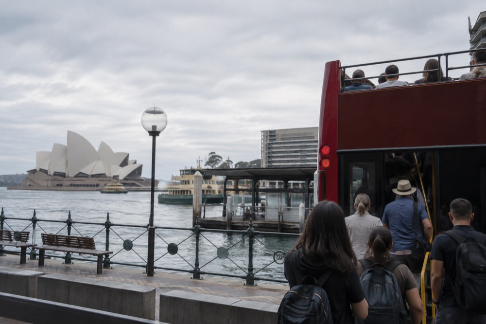 Passengers boarding an open-top bus at a city stop marked with a sightseeing sign