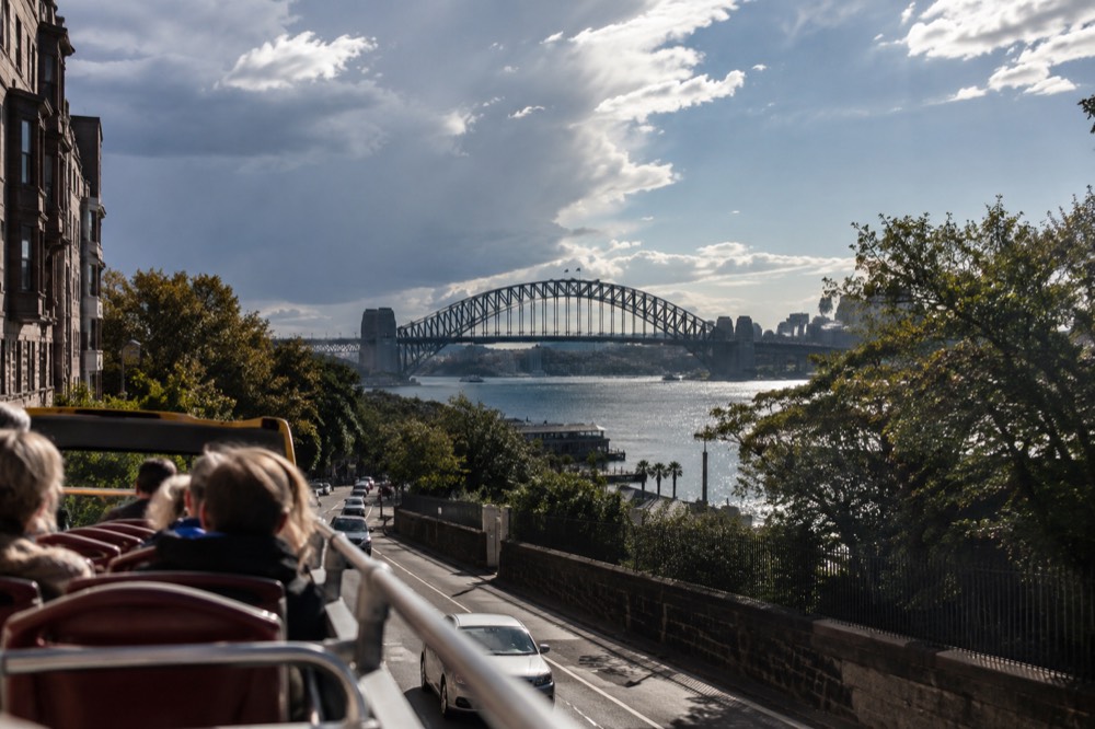Open top deck of a sightseeing bus with harbour bridge views along a city street