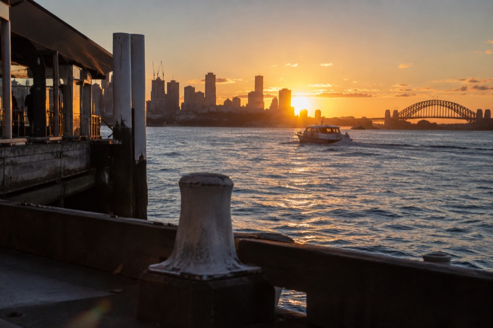 A small boat crossing the harbour at sunset with the city skyline glowing orange behind it