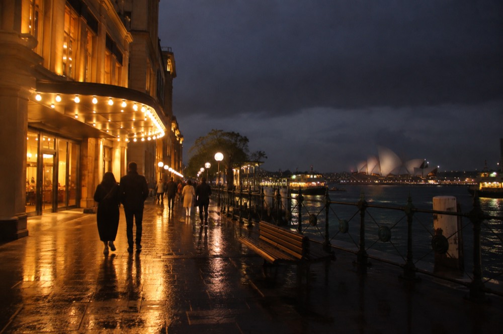 Warm bulb light spilling from a theatre entrance onto a wet city footpath at night