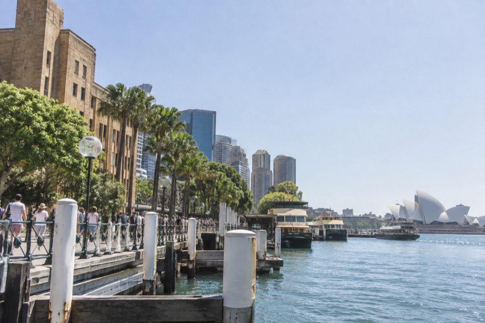Sunlit sandstone buildings along a Sydney harbour foreshore under a clear blue summer sky.