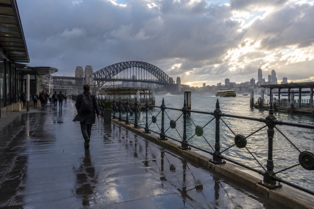 Rain clouds passing over Sydney Harbour, with the bridge and city towers partly lit by breaking sunlight.