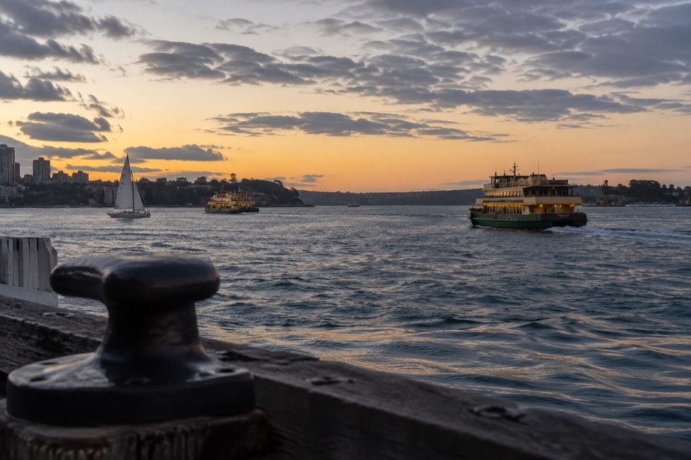 A sailing boat and ferries on Sydney Harbour under a partly cloudy sky between headlands.