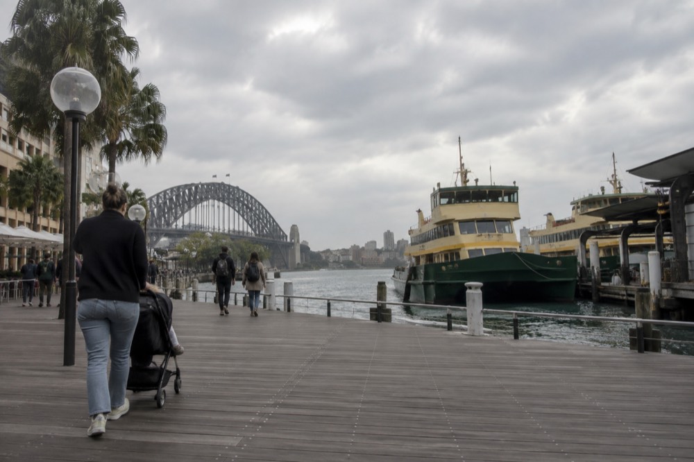 A stroller on a wide harbourside boardwalk with ferries moored across the water