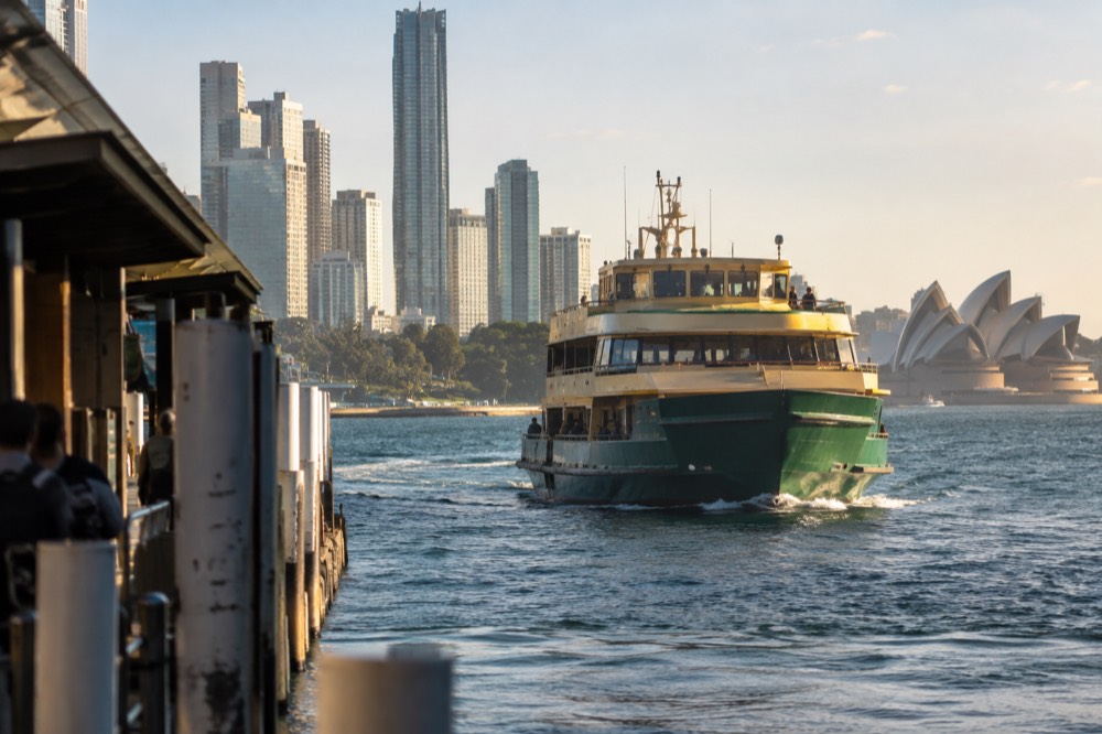 A green and yellow Sydney ferry approaching a wharf with the city skyline behind it