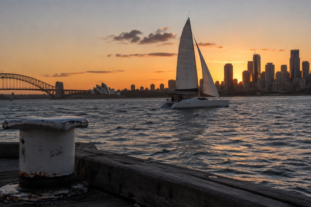 A catamaran under sail on Sydney Harbour at sunset with the city skyline glowing orange behind it