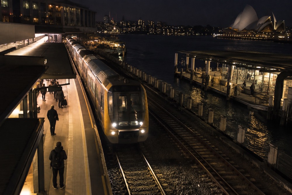 A lit train platform at night with a Sydney train arriving and a few passengers waiting