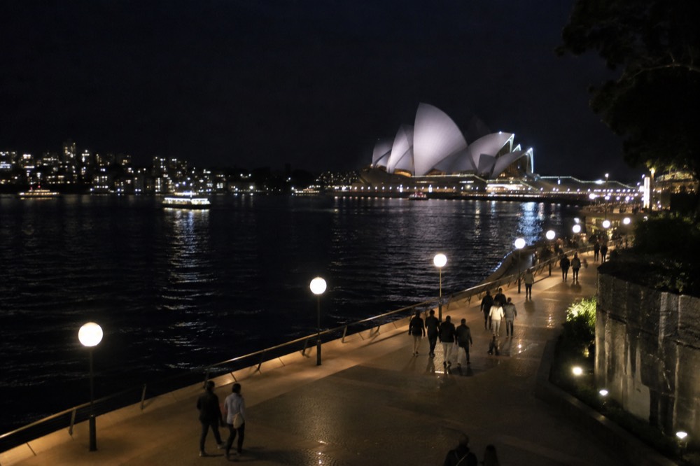 People walking a lit harbourside promenade at night with the Opera House lit in the background
