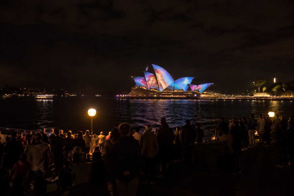 Crowds gathered on Circular Quay foreshore watching colourful light projections on the Opera House sails at night