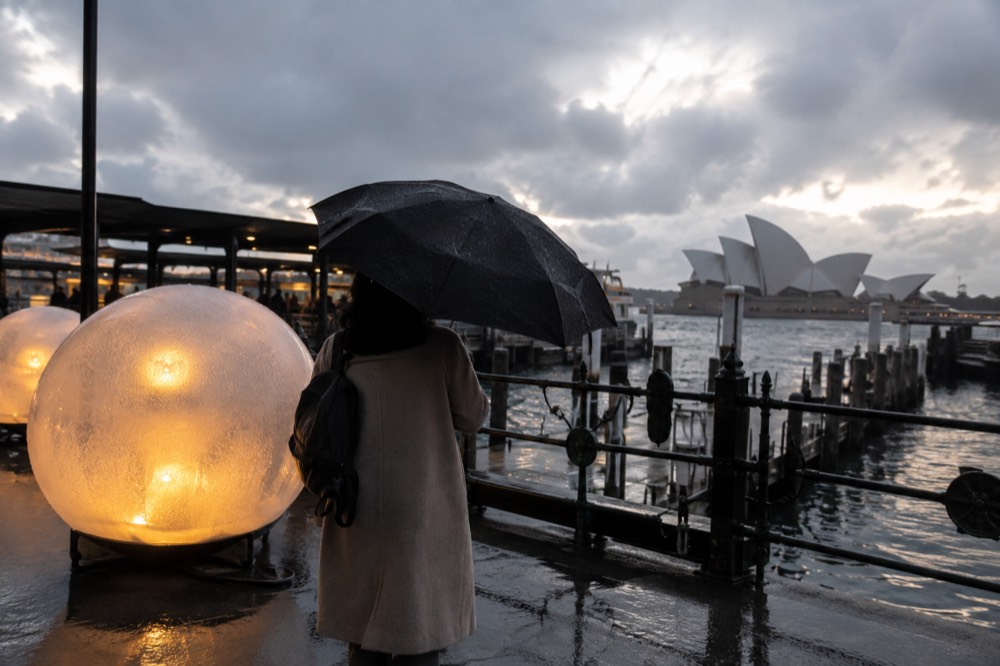 Person in a warm coat holding an umbrella beside a glowing light installation in light rain