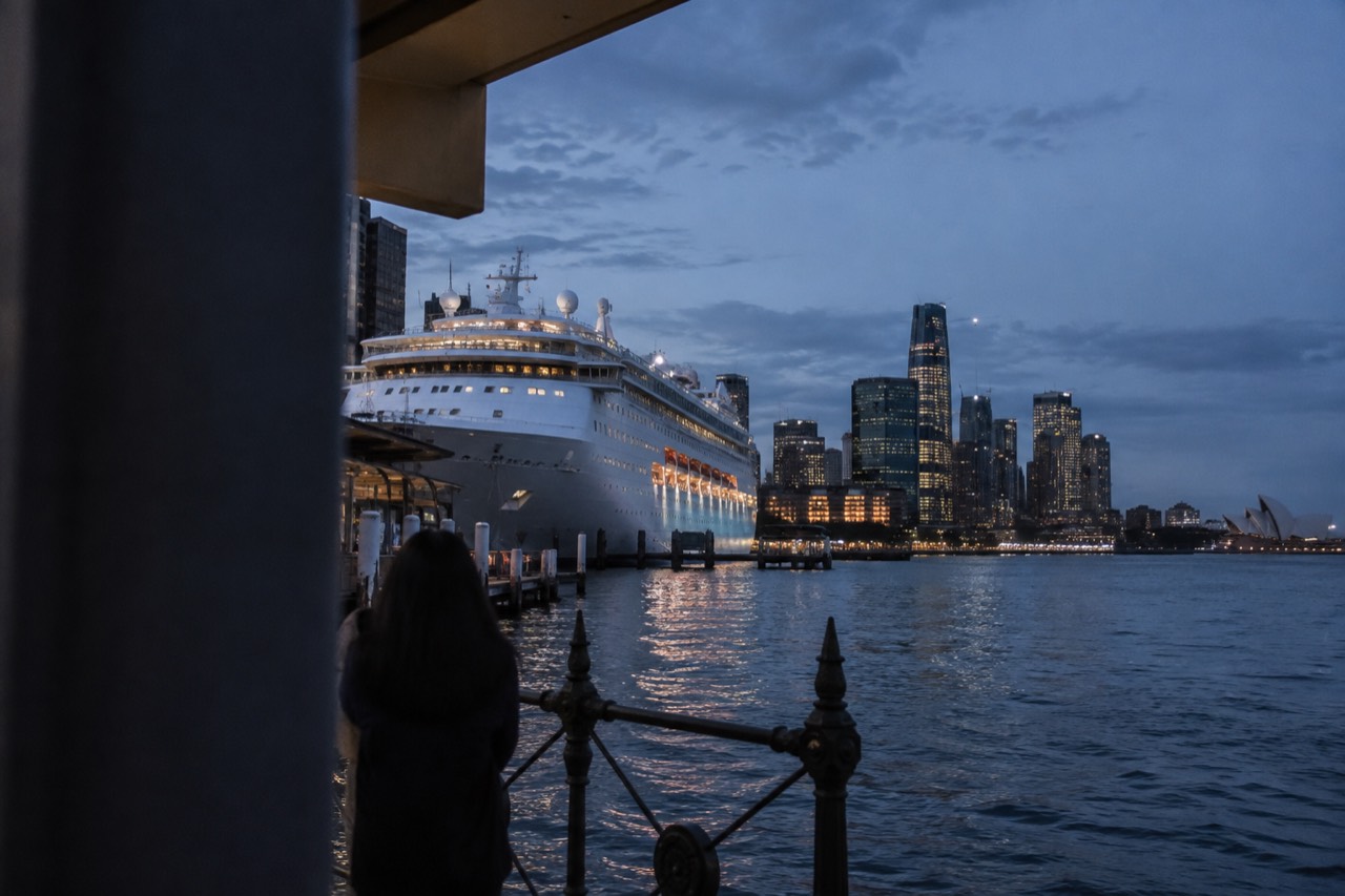 A white cruise ship docked at a Sydney harbour terminal with the city skyline behind it